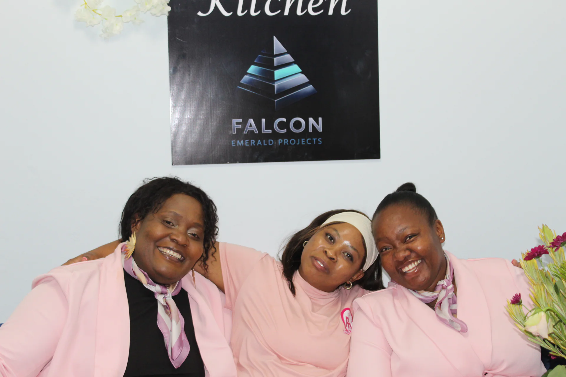 Three Falcon Kitchen team members in pink attire seated below branded Falcon Kitchen signage